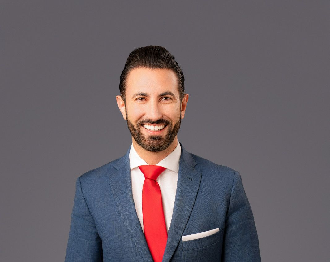 A professional portrait of a man in a blue textured suit, white shirt, and red tie, smiling against a grey background.