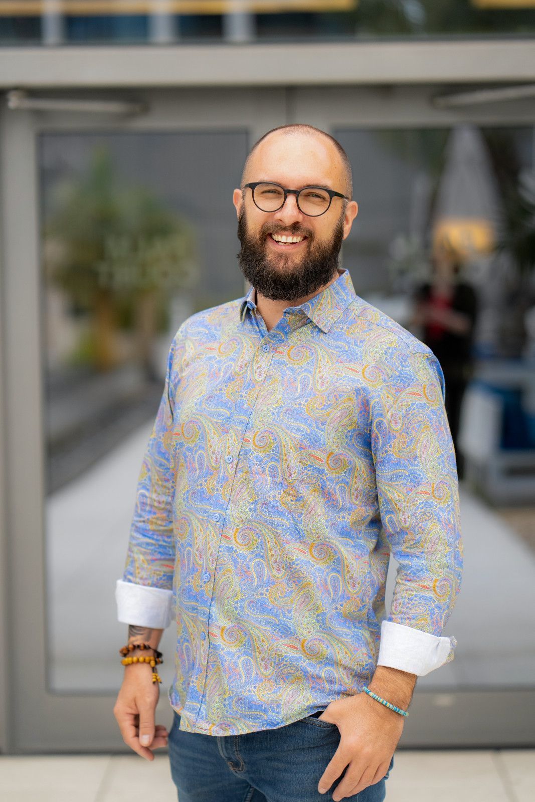 Smiling man with glasses in a blue patterned shirt, standing outdoors in front of glass doors