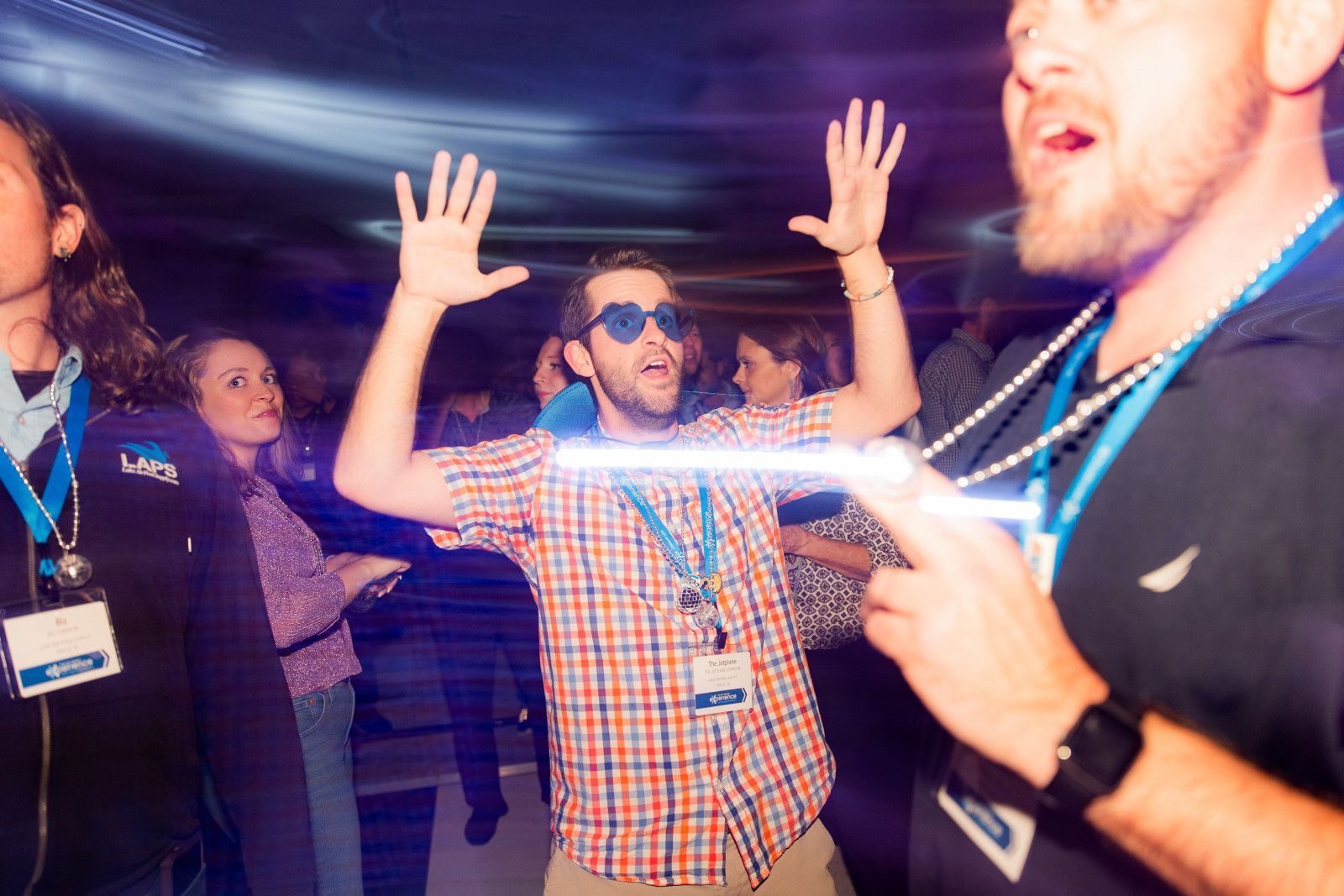 conference photographer orange county People in a dark venue with colorful lights, including a person with sunglasses and hands raised in the air at conference after party in orange county