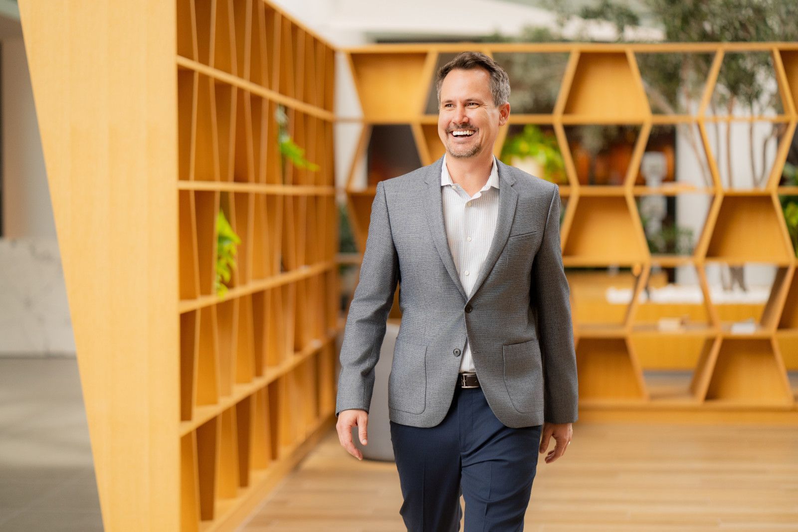 Man in gray blazer walking through a modern office with wooden shelves, smiling for financial advisor office in Irvine