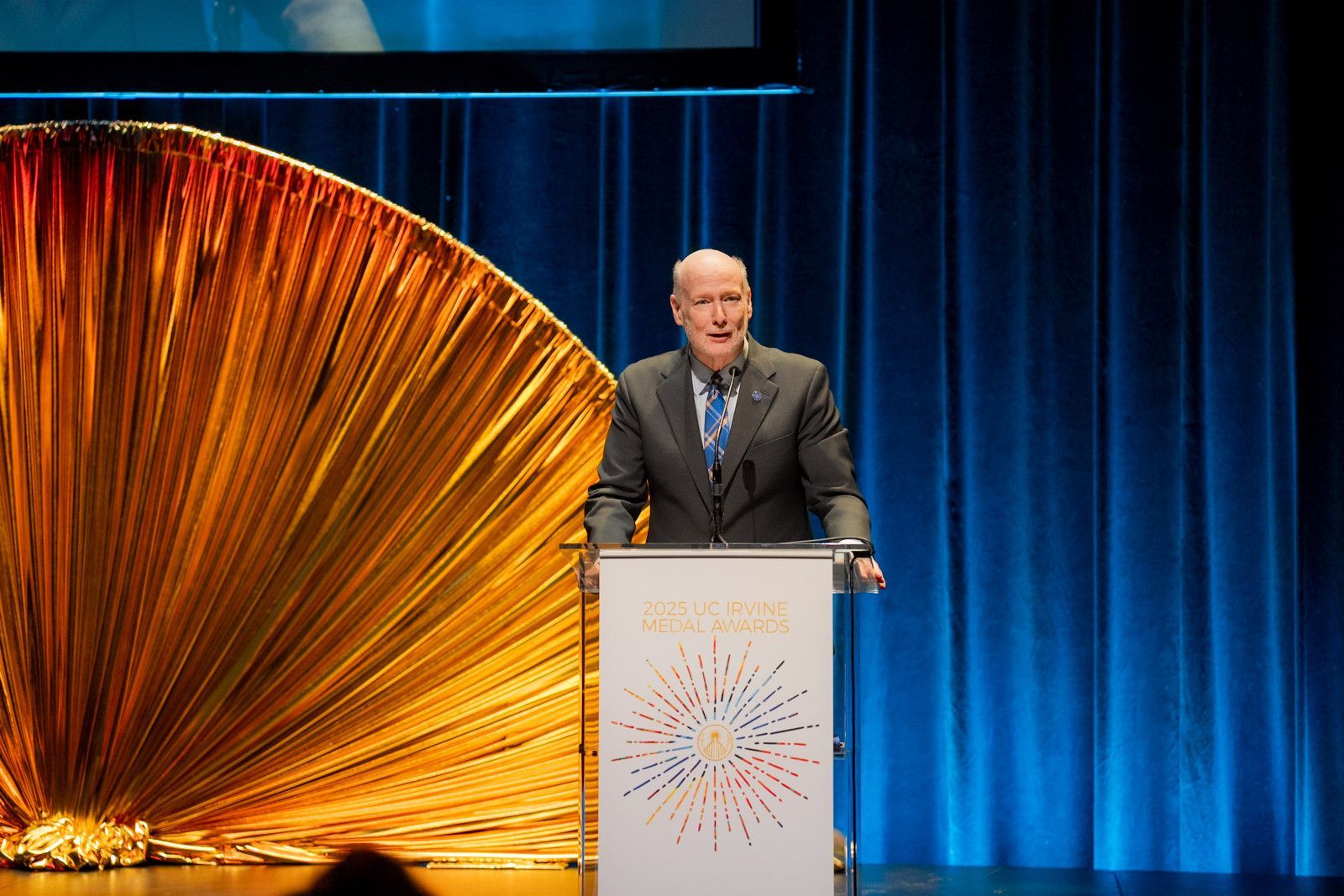 Chancellor speaking at a podium on a blue-lit stage beside a large orange fan-like backdrop at UCI event