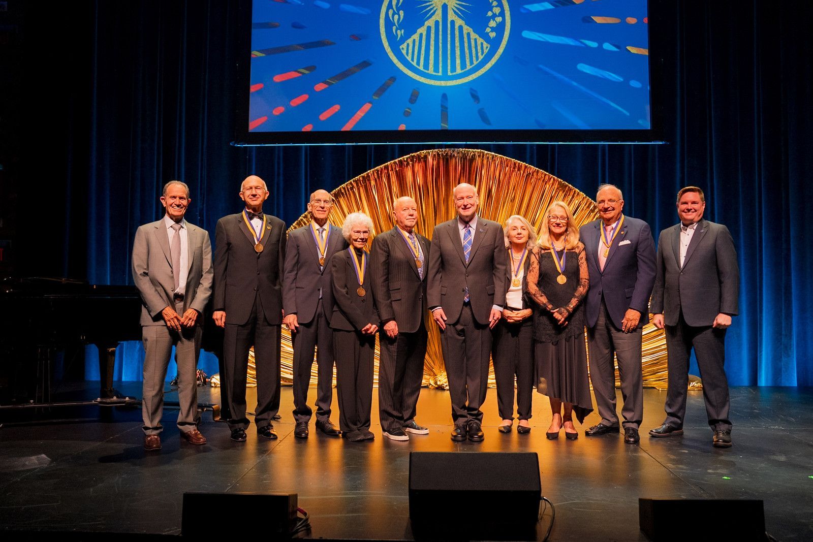 Group of people standing on a stage under blue lighting, with a large screen and circular logo behind them for UCI donor event