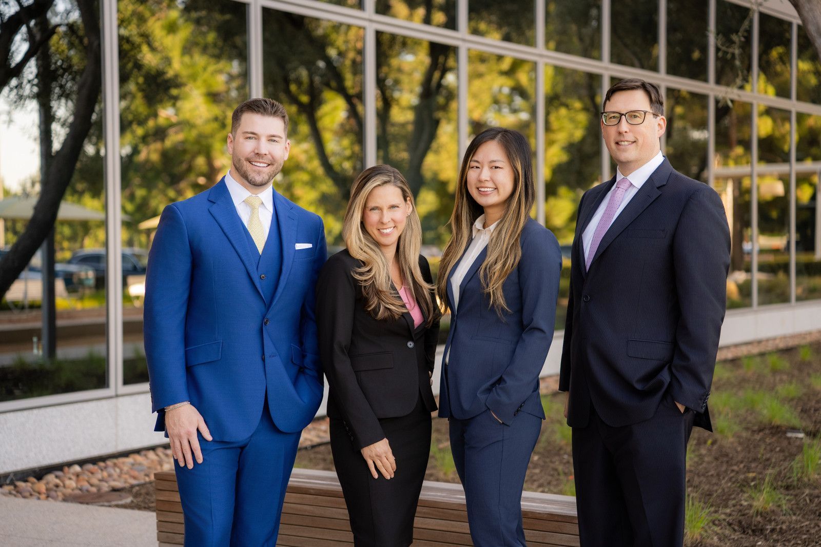 orange County Law Firm Four professionals in business attire stand smiling together outside a modern office building with large glass windows.