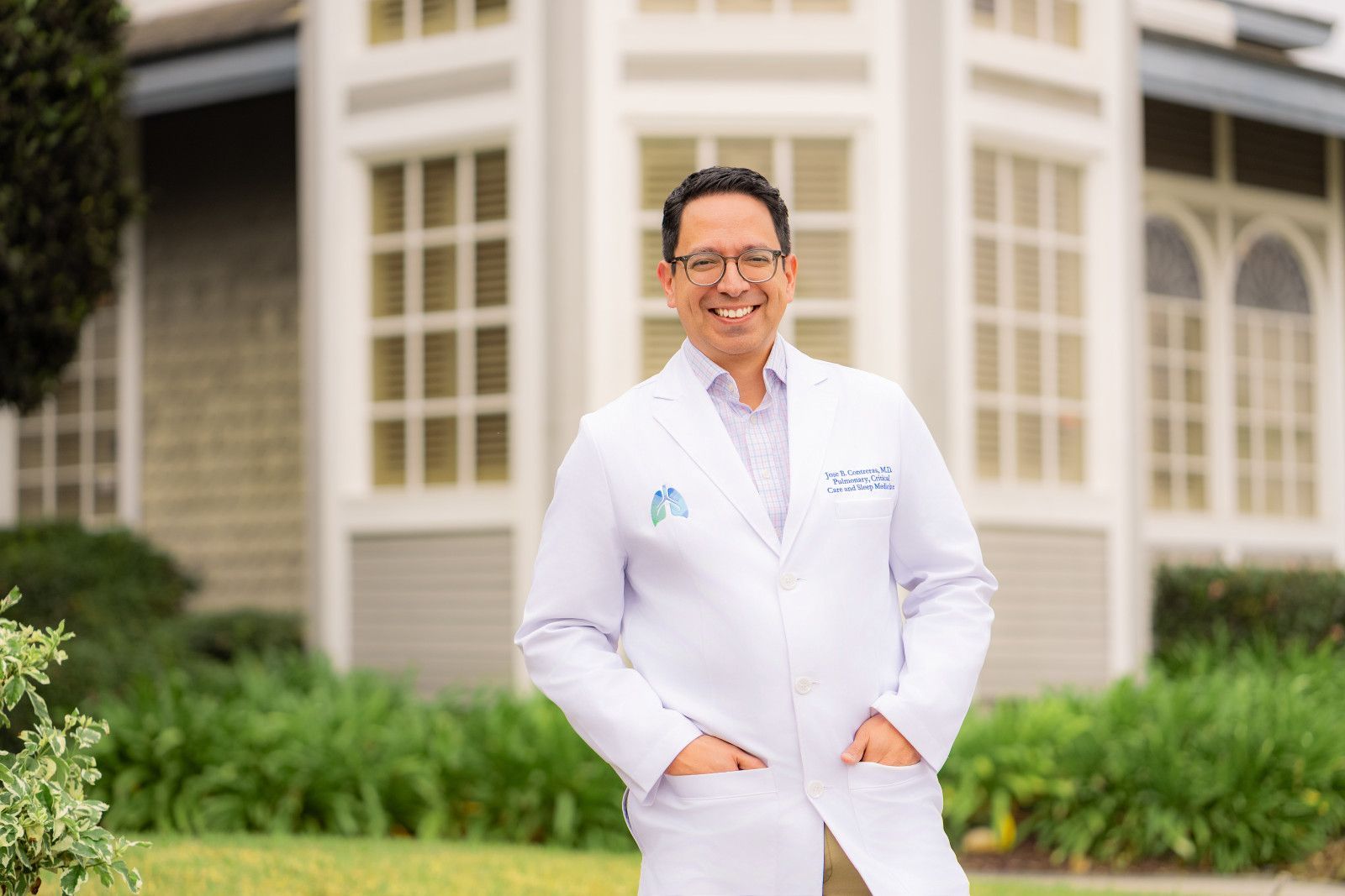 Smiling doctor in a white coat standing outside a building with large windows and greenery in Orange County, CA