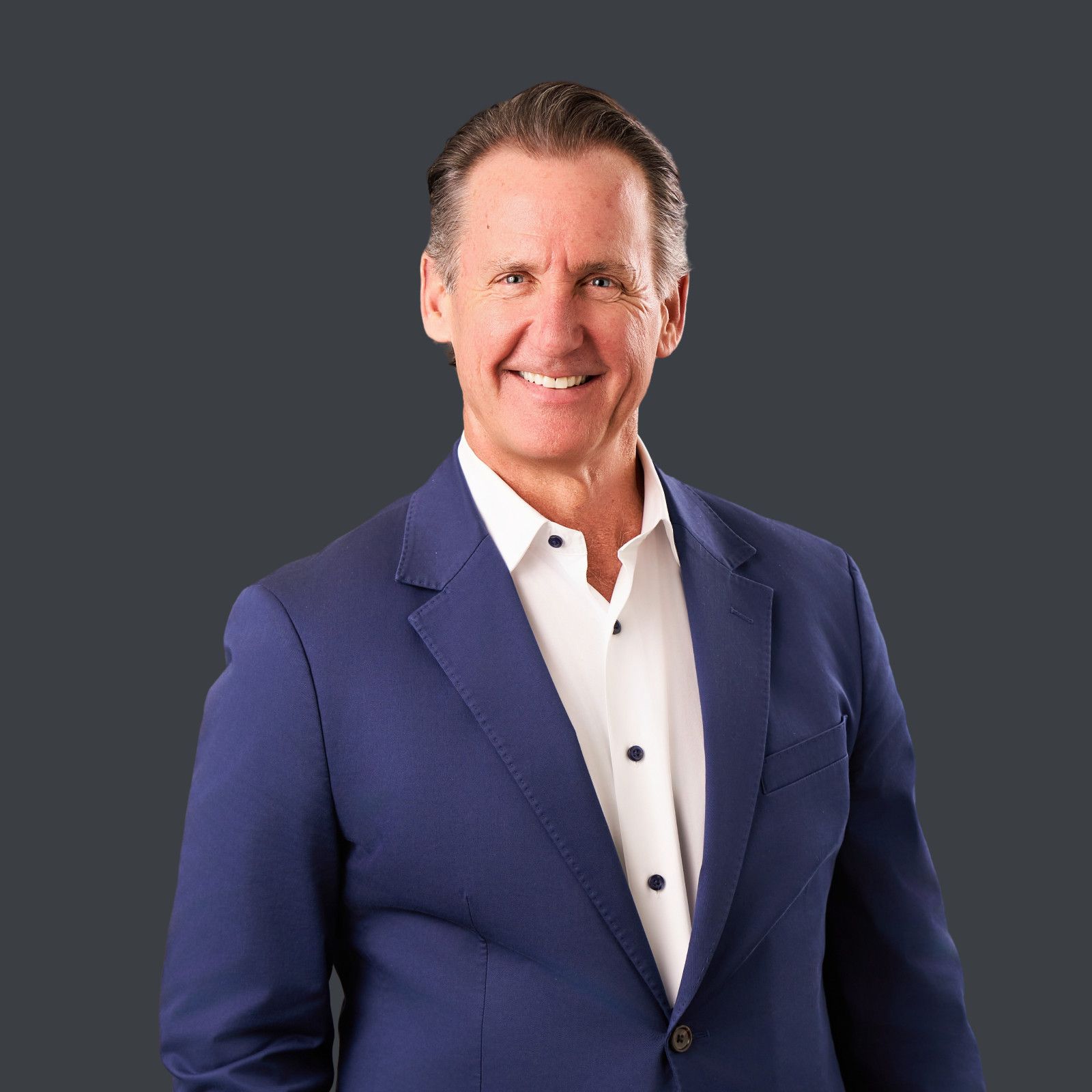 Newport Beach Male Executive Headshot Man in dark blue blazer and white shirt smiles, hands in pockets against gray backdrop in Newport Beach office