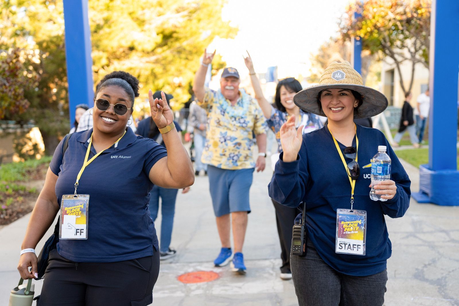 Two smiling walkers wave on a sunny path, wearing navy shirts and lanyards at an outdoor event at UCI campus