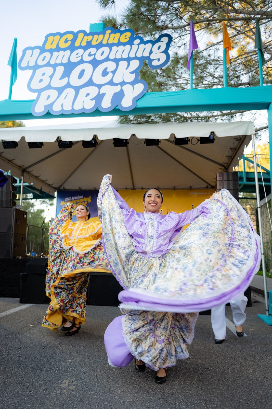 Dancers in yellow and lavender costumes perform under a “Block Party” sign at an outdoor event for UCI homecoming