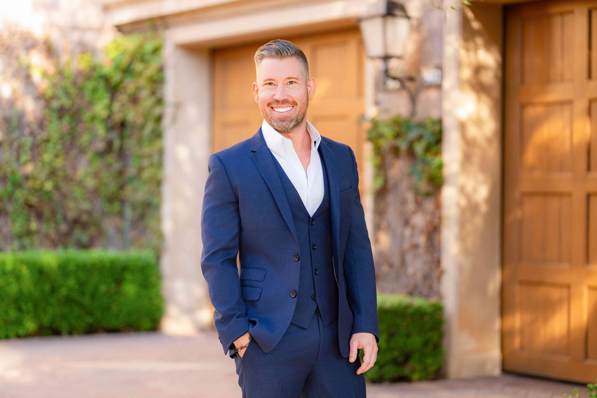 Man in navy suit smiling outdoors beside a building with stone columns and wooden doors, realtor photo
