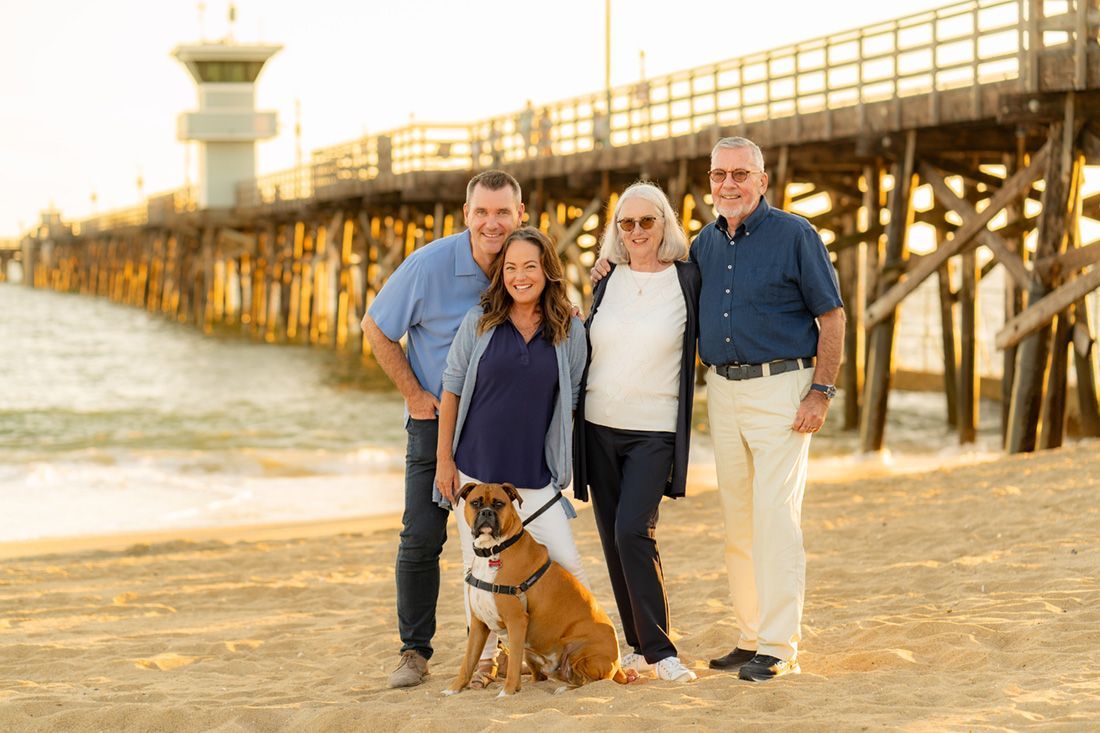 A group of people and a dog are posing for a picture on the beach in front of the Balboa Pier. 