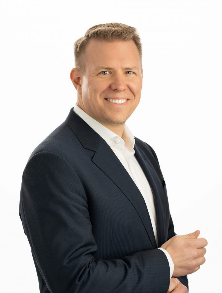 A man in a suit and white shirt is smiling for the camera in front of a bright white backdrop 