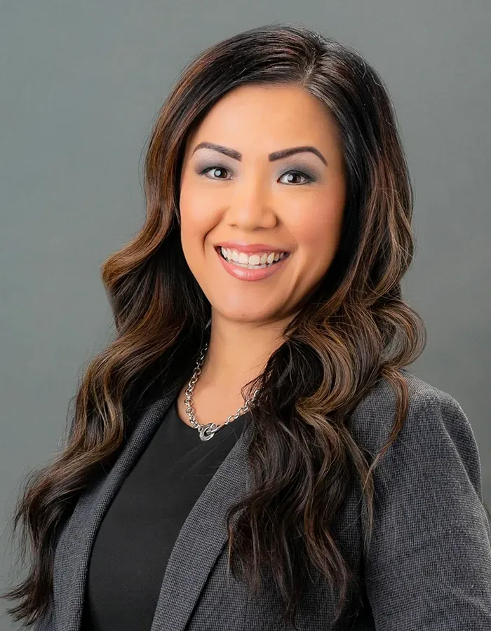 A woman with long hair is wearing a gray jacket and smiling.