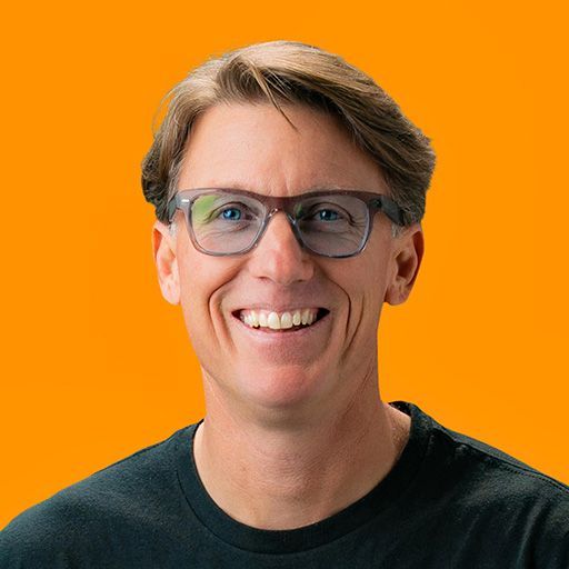 Christopher Todd wearing glasses and a black shirt is smiling in front of an orange background.