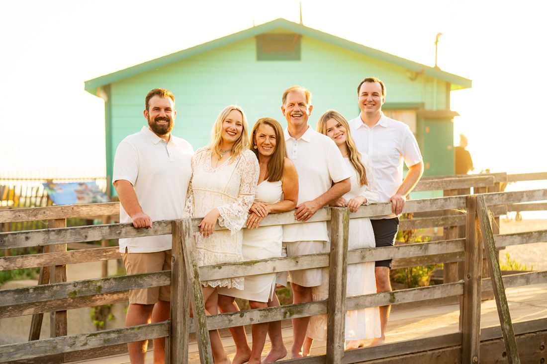 Family posing near a weathered wooden fence with a light green building in the background. They are wearing white.