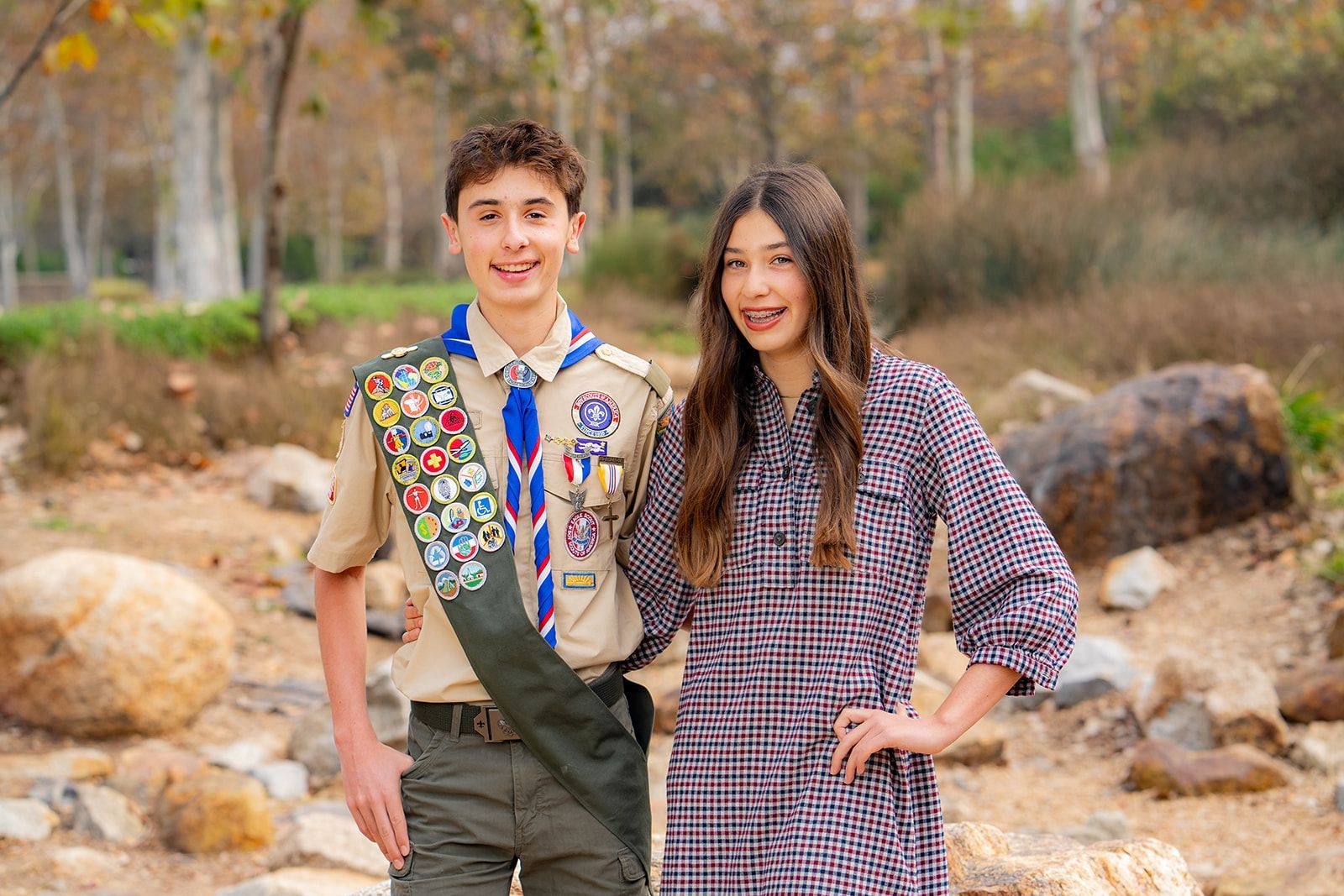 Boy in Scout uniform stands with girl in plaid dress, both smiling outdoors at park in Irvine. 
