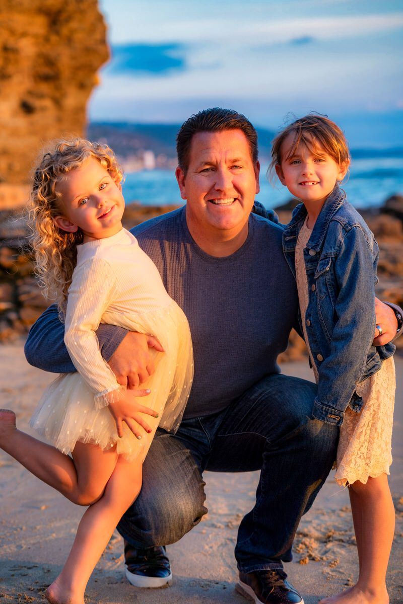 A dad is kneeling with his young daughters on a beach.