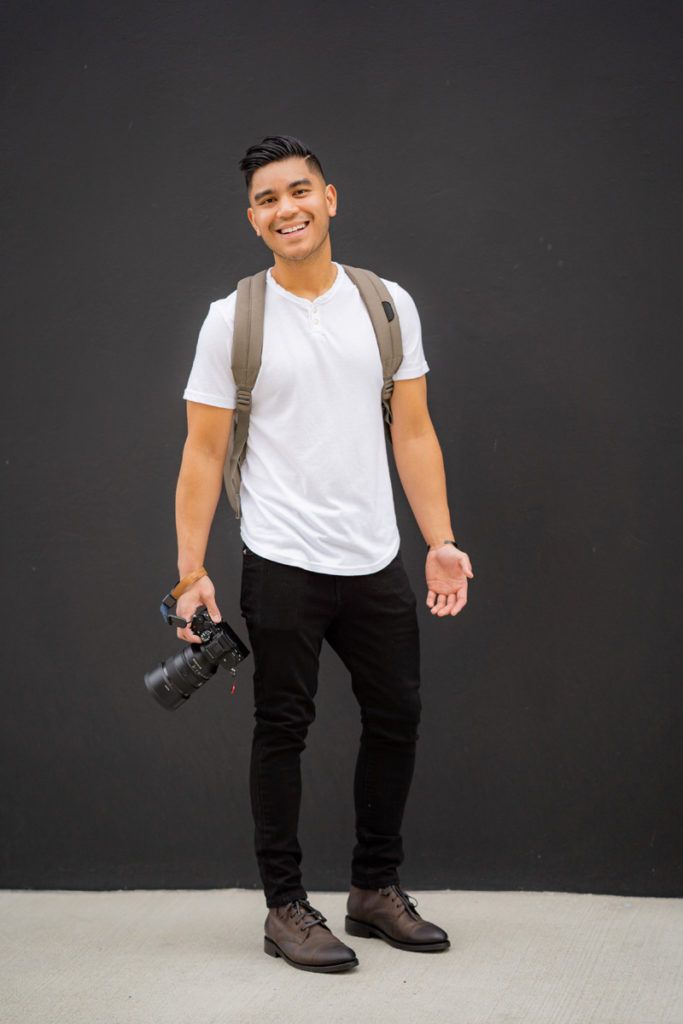 A young man is standing in front of a black wall holding a camera for a dating photoshoot