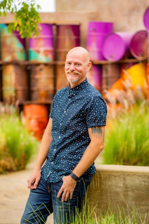 A man during a dating photoshoot is leaning against a wall in front of a wall of barrels.