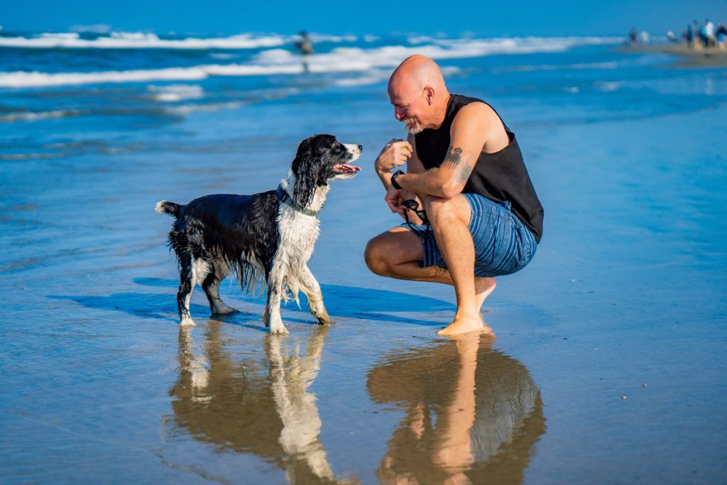 A man is kneeling down next to a dog on the beach during a dating photo shoot