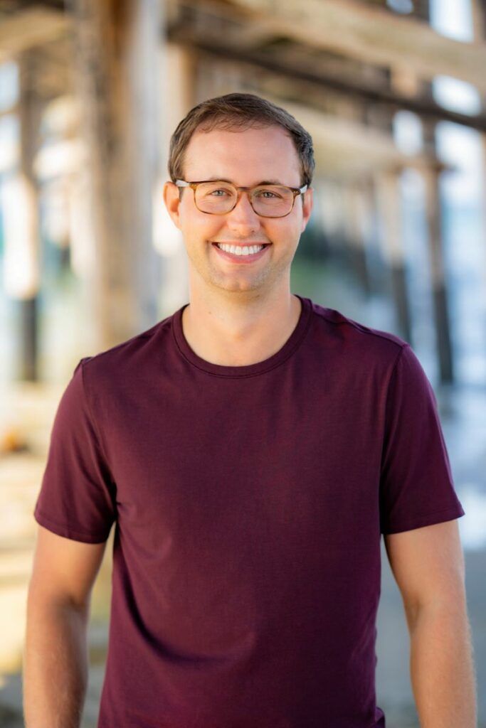 An Actor wearing glasses and a purple shirt is smiling for the camera.