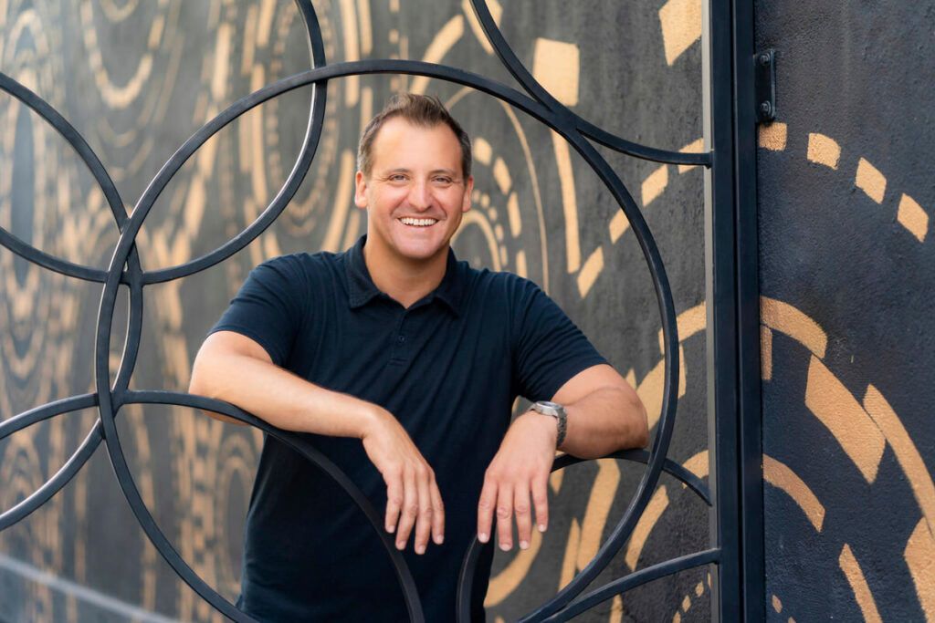 A man in a black shirt is leaning against a metal fence and smiling.
