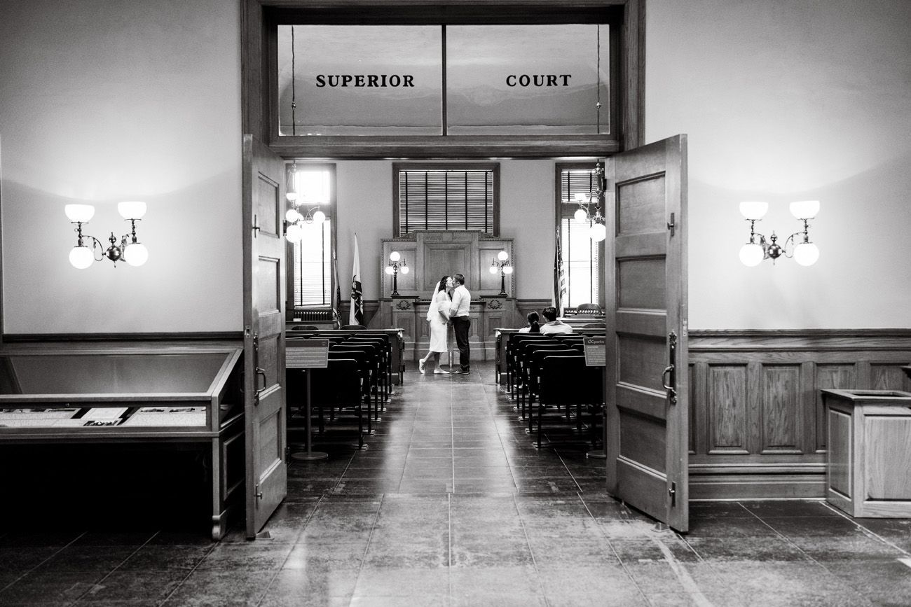 A black and white photo of a bride and her parents kissing her on the cheek in the Historic Courtroom in Santa Ana. 