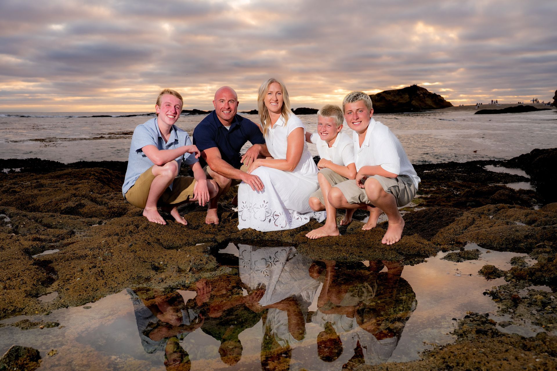 Family of five crouches on rocks, ocean backdrop. Sky with orange hues reflects in water at Treasure island beach in laguna