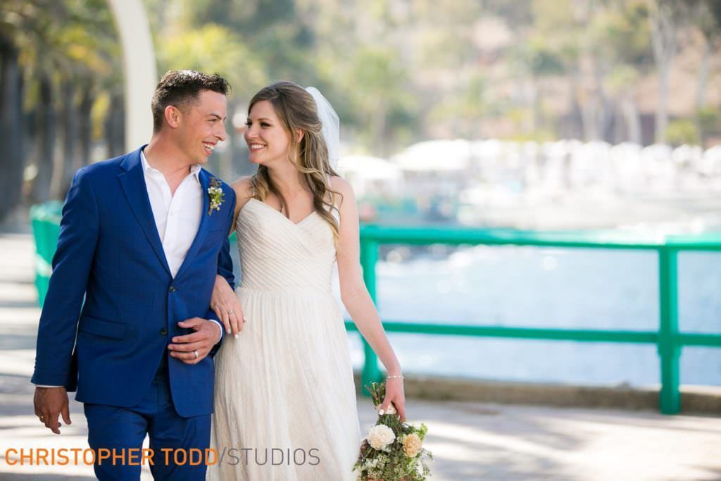 A bride and groom are posing for a picture in front of Avalon Harbor in Catalina