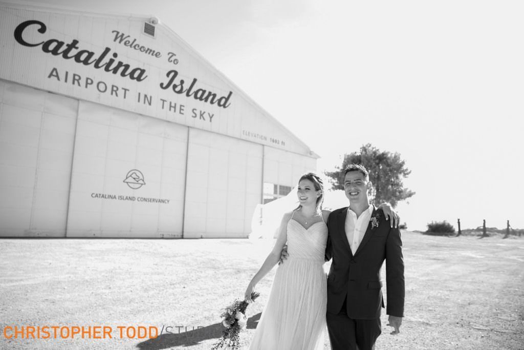 A bride and groom are walking in front of a sign that says welcome to catalina island airport in the sky
