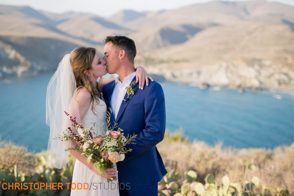 A bride and groom are kissing in front of shark Harbor in Catalina