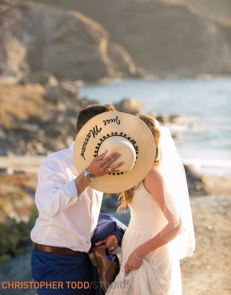 A bride and groom kissing on Catalina Island while the bride covers her face with a hat.