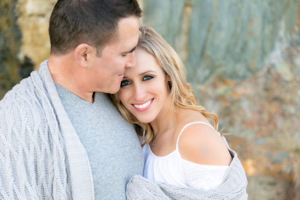 An engaged man is kissing his fiancée on the cheek while wrapped in a blanket.