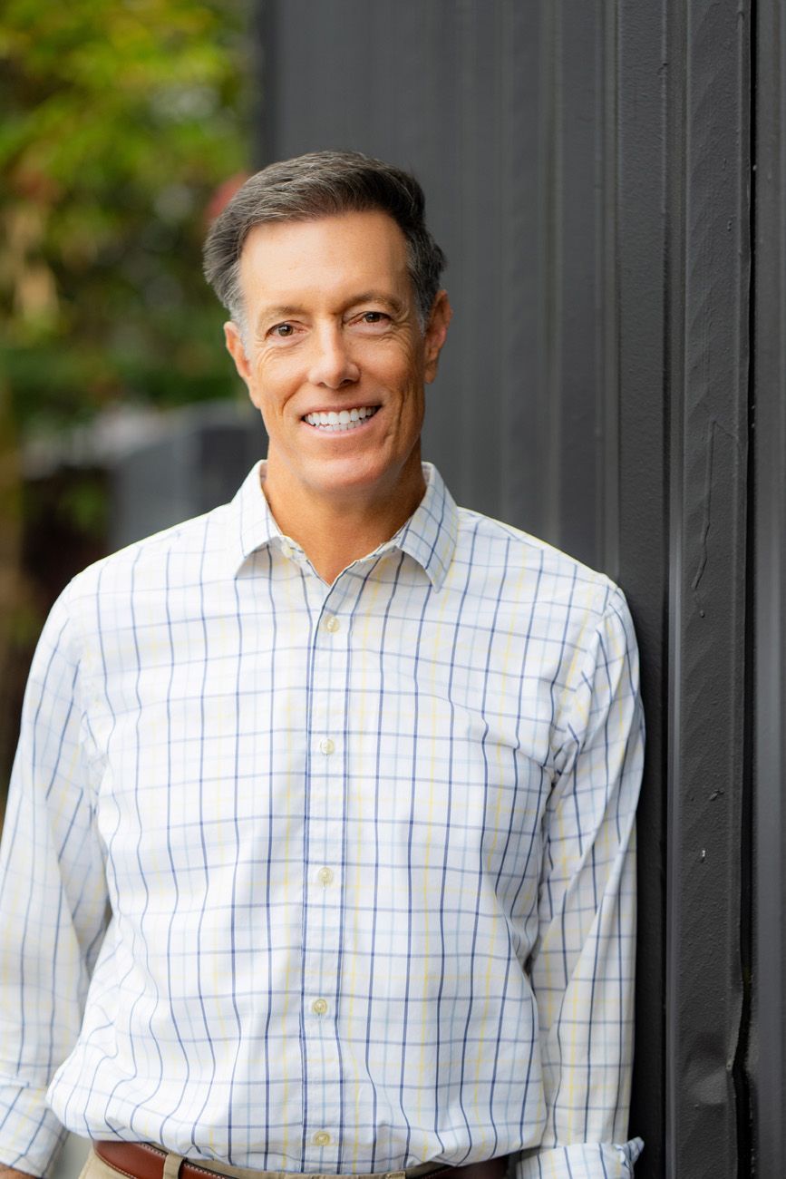 Smiling man in a plaid shirt standing by a dark wall indoors for costa mesa realtor headshot