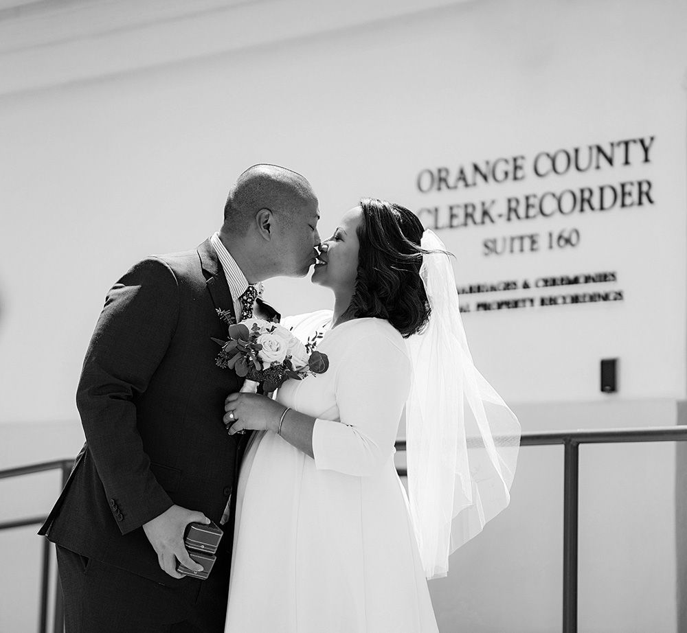 A black and white photo of a bride and groom kissing in front of an orange county clerk recorder sign.