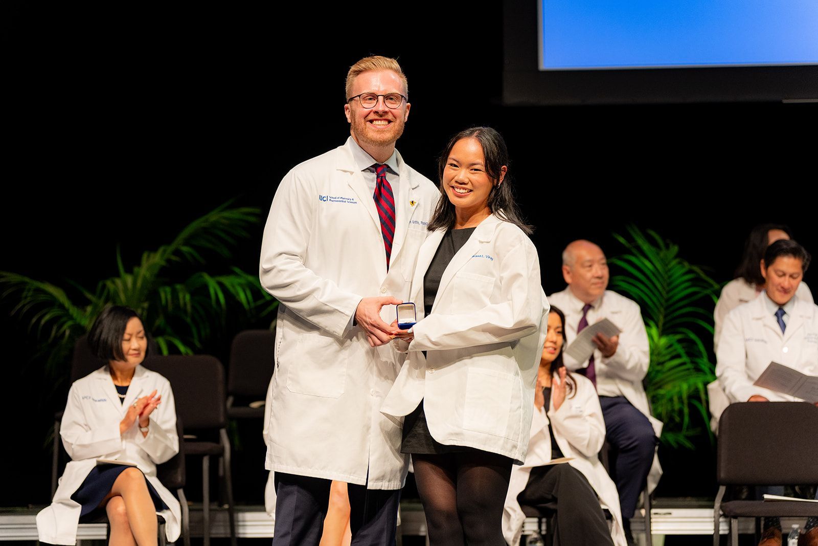 Two people in white coats smiling on stage, shaking hands during a white coat ceremony at UCI