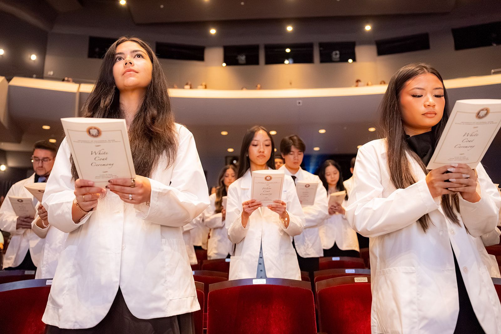 People in white blazers stand in an auditorium, taking their oath for pharmacy students at UCI holding certificates during a ceremony.