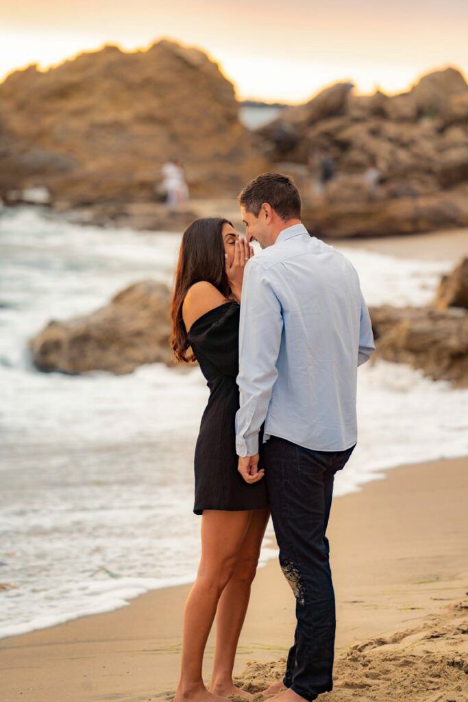 A man and a woman are kissing on the beach after a surprise engagement
