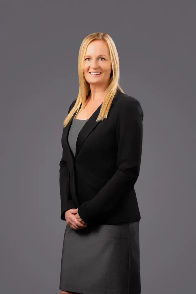 A woman in a black jacket and gray skirt is standing in front of a gray background.
