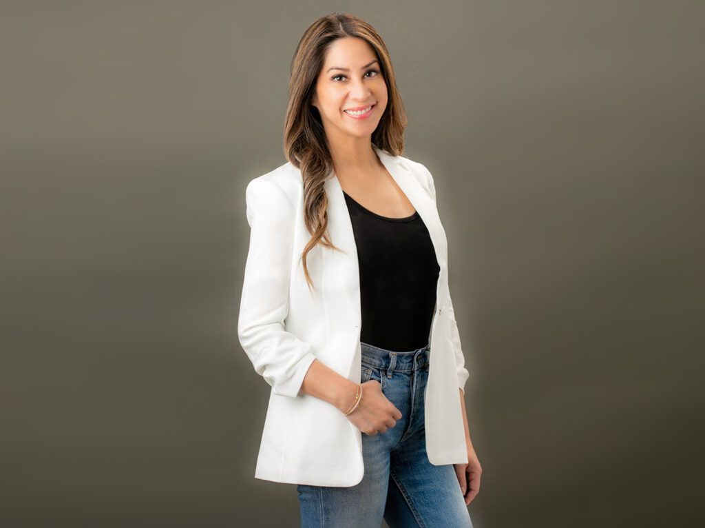 A woman wearing a white jacket and jeans is standing in front of a dark gray wall.