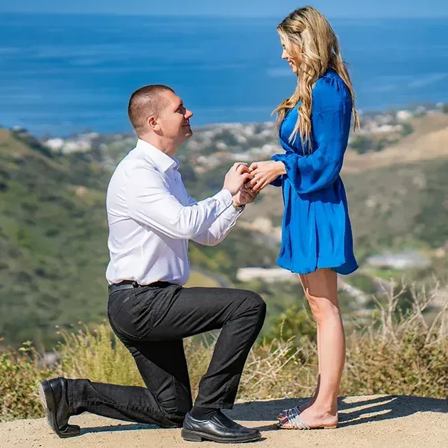 A man is kneeling down to propose to a woman in a blue dress at Top of the world park  in Laguna Beach