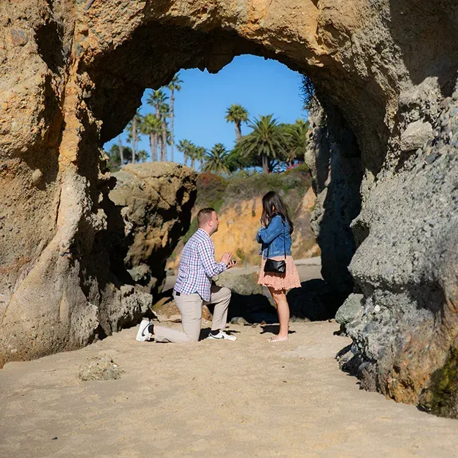 A man is kneeling down to propose to a woman at the Keyhole at Treasure Island Beach in Laguna Beach 
