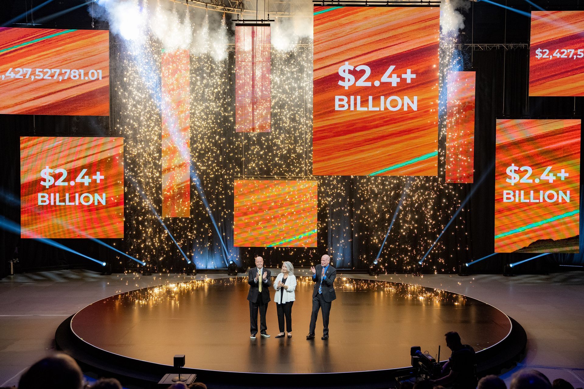 Three presenters on stage beneath large orange screens reading “$2.4+ BILLION” as confetti falls