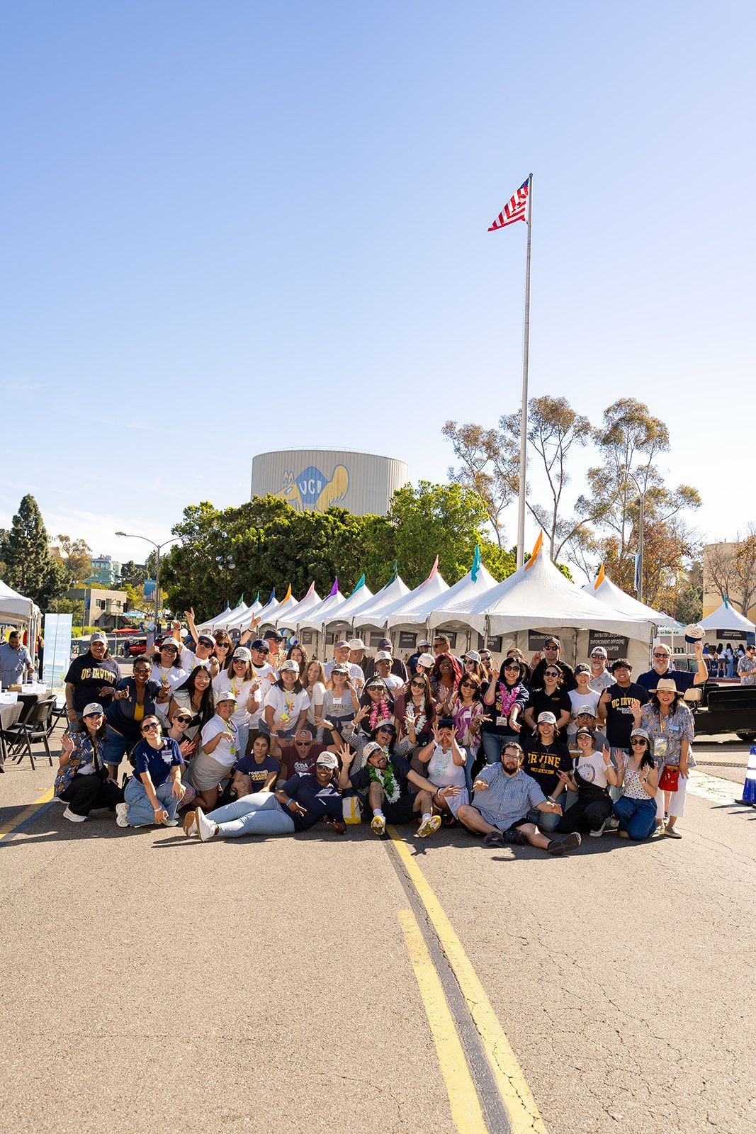 Large group posing on a sunny street with white tents and a flag in the background on UCI campus