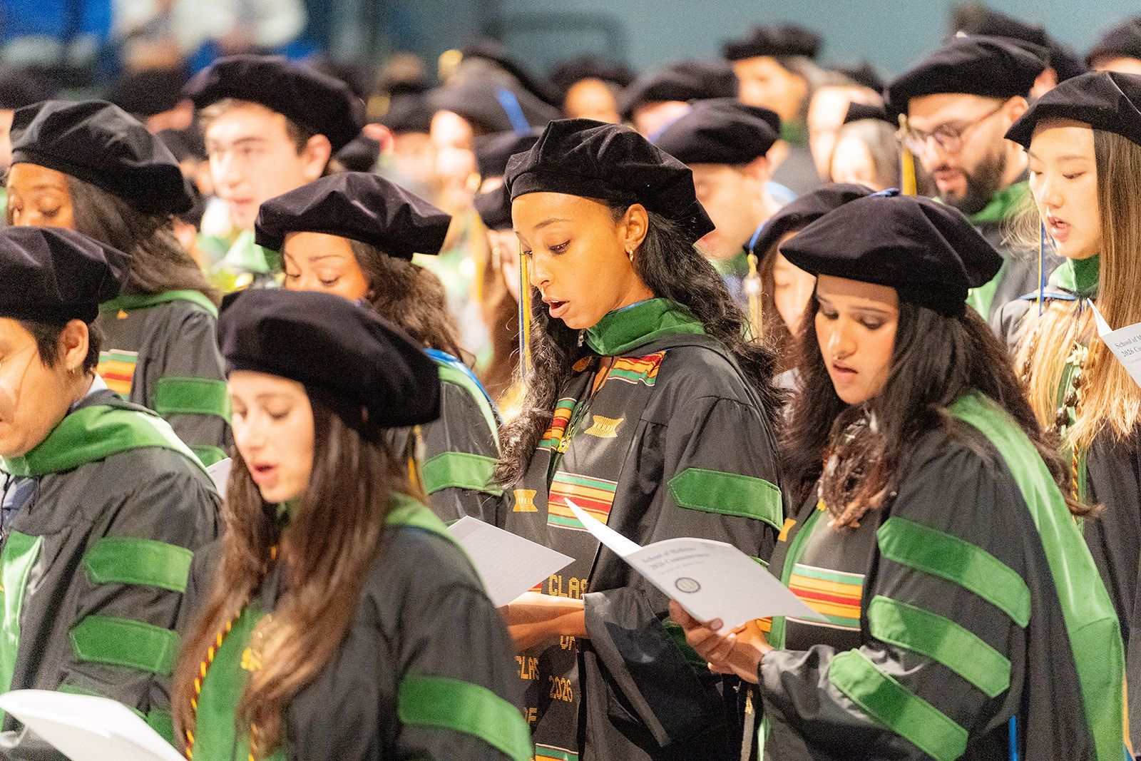 Graduates in black caps and green-trimmed gowns reading programs at a ceremony for medical students at UCI