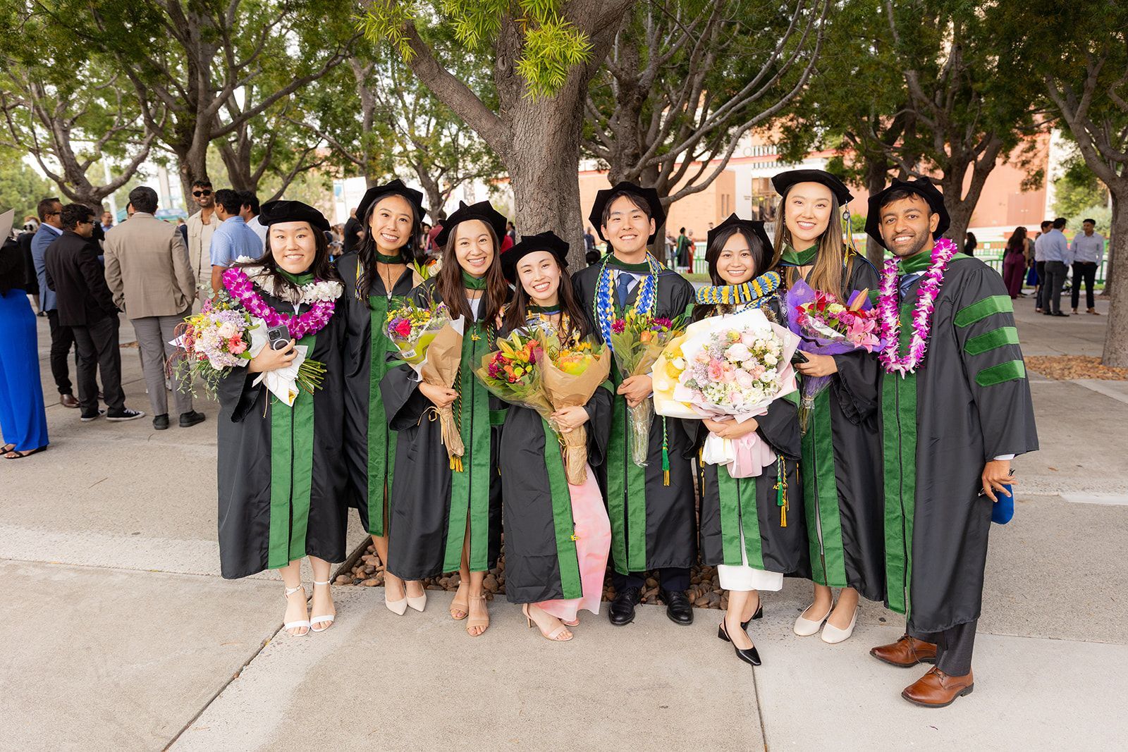 Graduation group posing outdoors in caps and gowns, holding bouquets and leis under trees for school of medicine grads at UCI