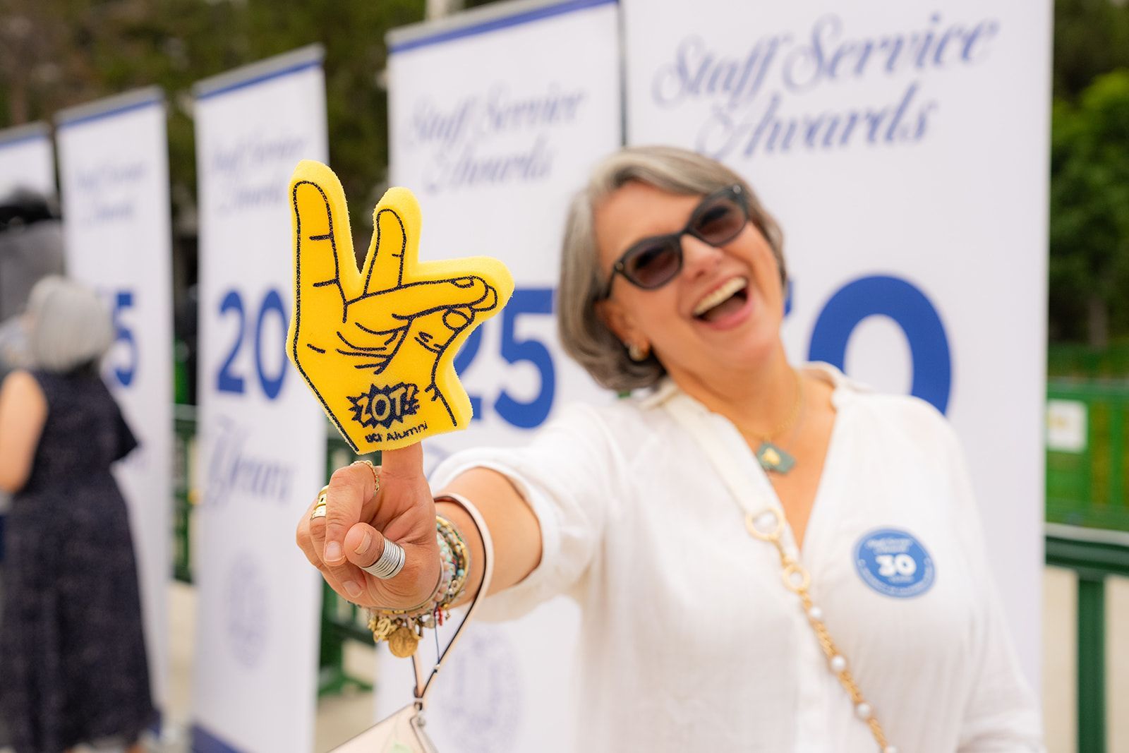 Woman in white outfit smiling and holding up a yellow foam hand at an outdoor awards event