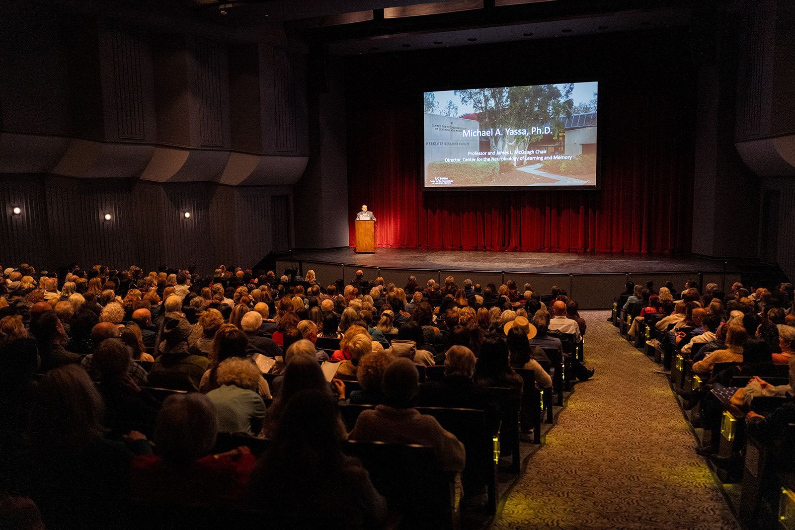 Audience watches a presentation in a theater, with a speaker on stage and a large screen above red curtains at UCI auditorium