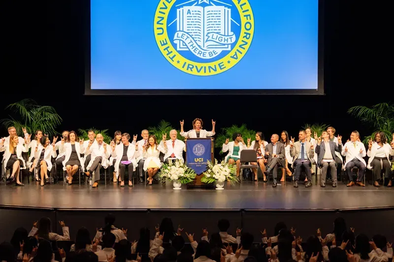 Graduation ceremony on stage with seated graduates, podium, and University of California Irvine seal projected above