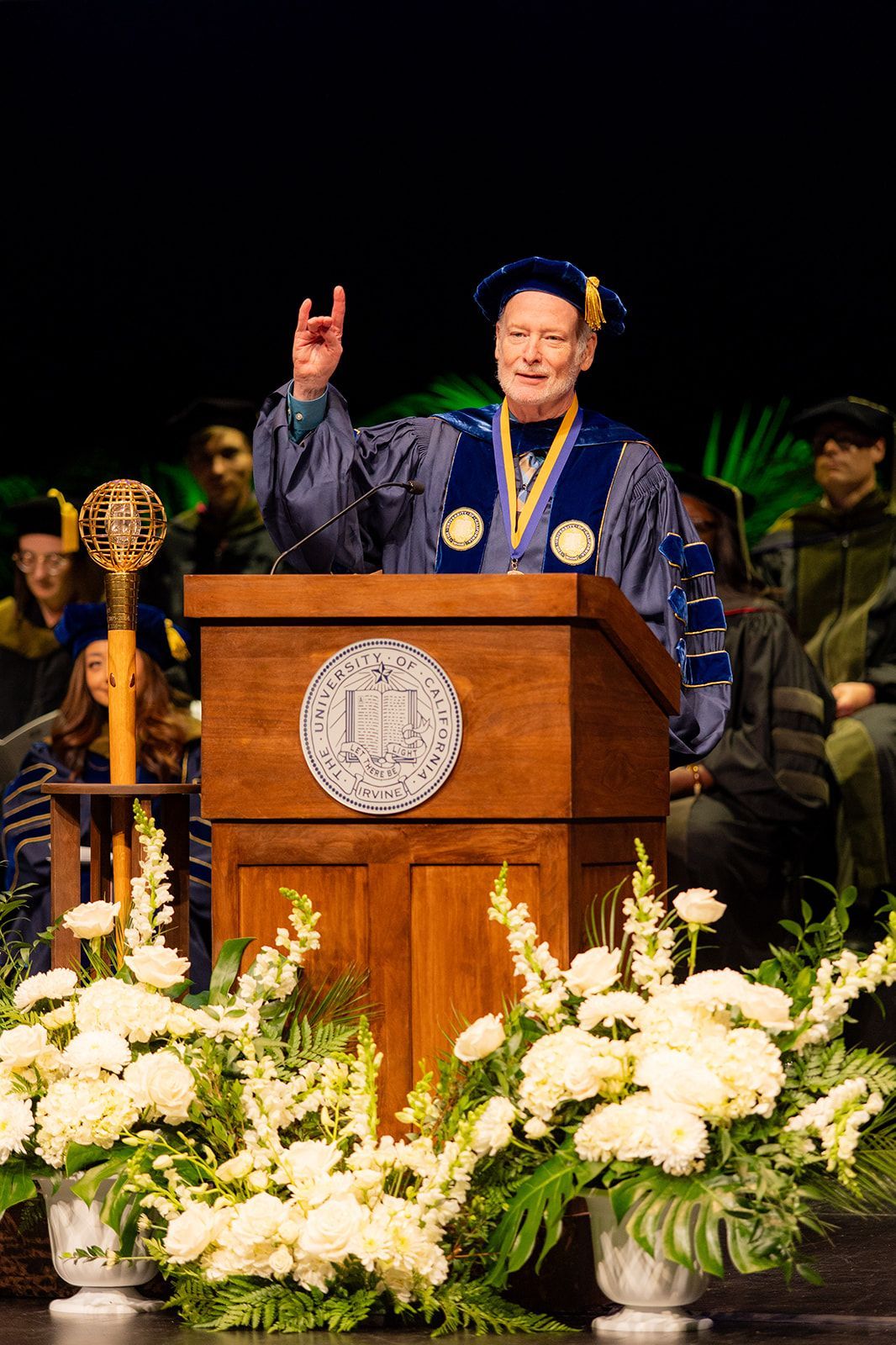 Speaker at podium on stage, wearing blue academic regalia and cap, surrounded by white flowers