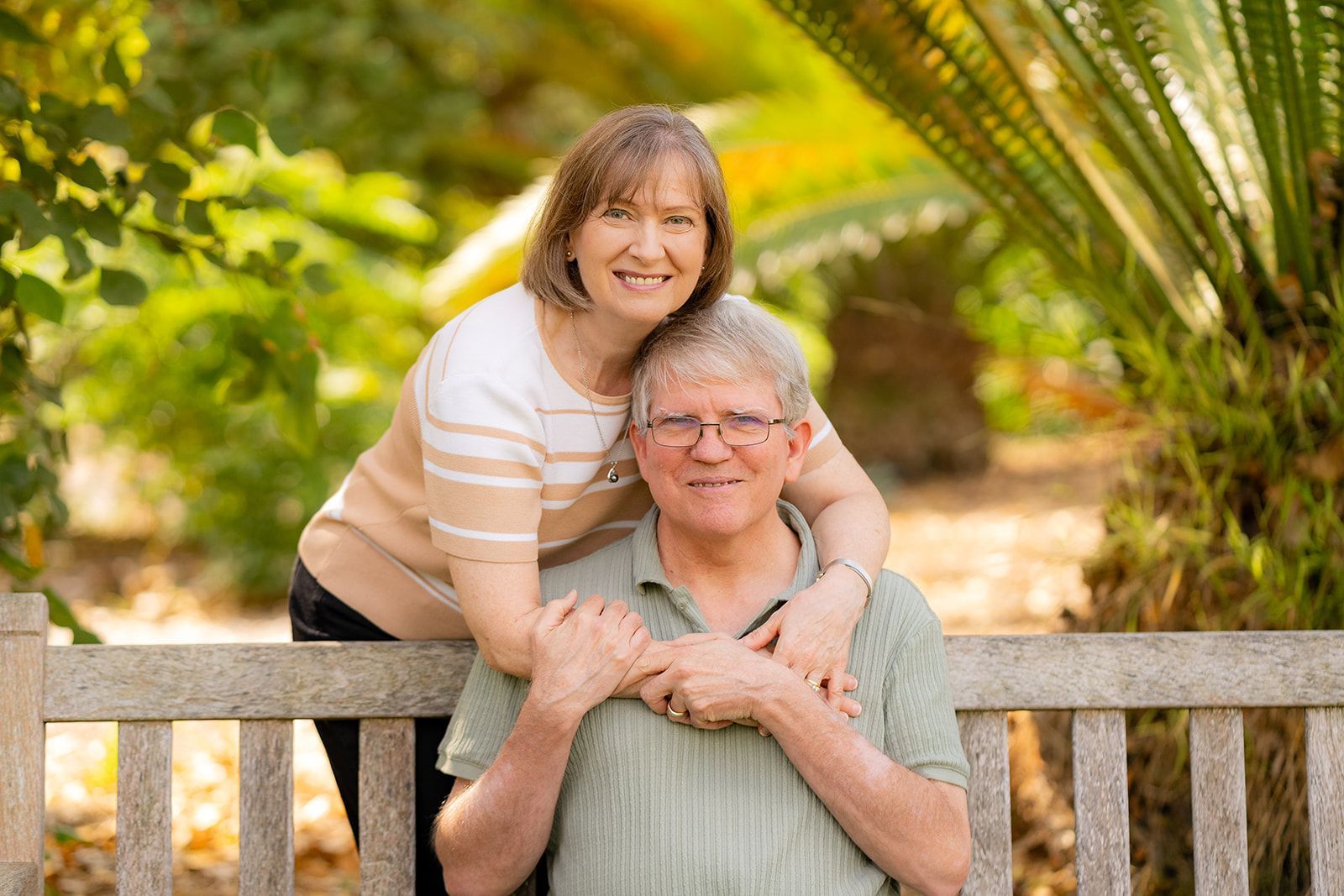 Woman embraces man sitting on a wooden bench outdoors; both smiling, greenery in background at Fullerton Arboretum