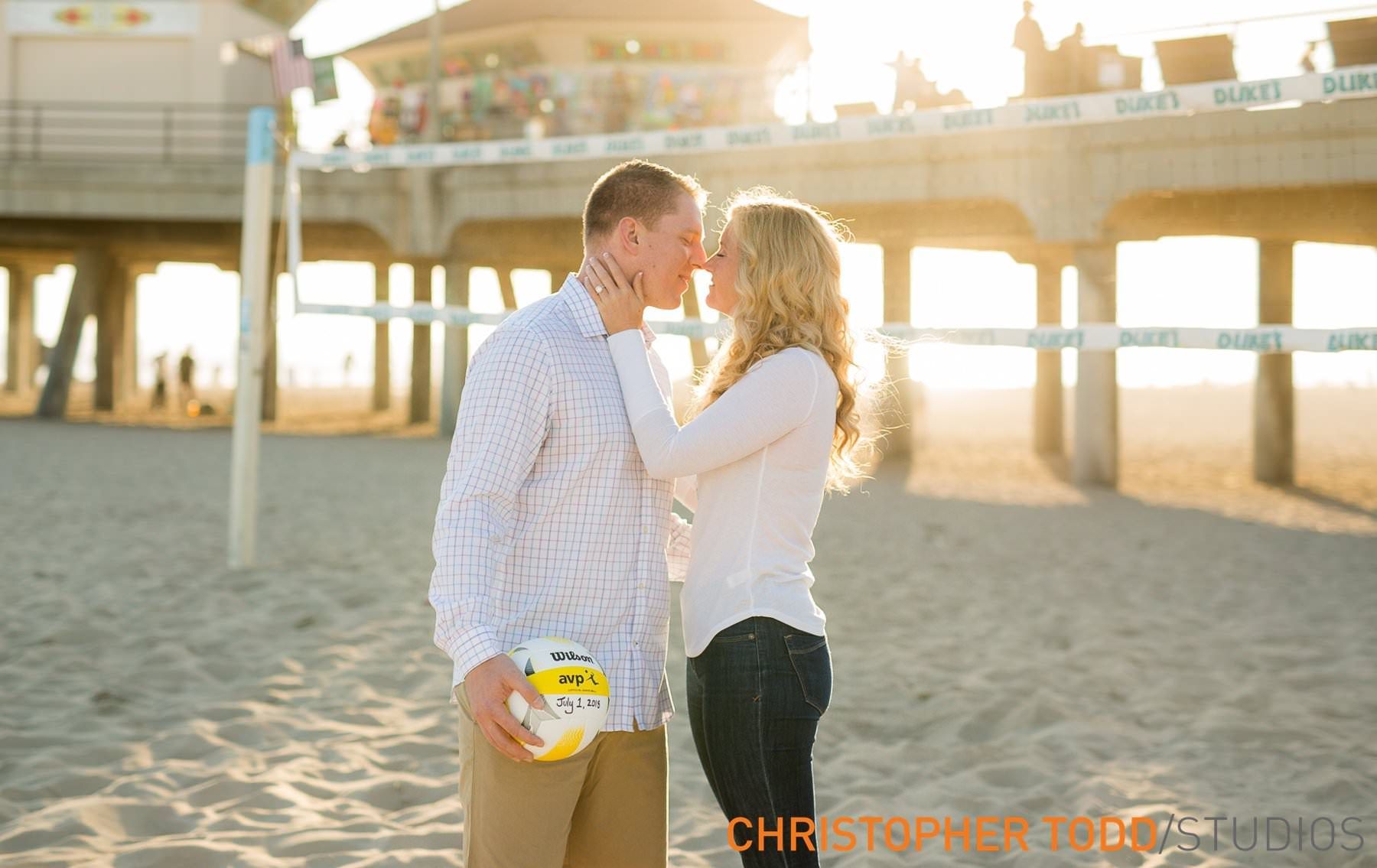 A man and a woman are kissing on the beach while holding a volleyball at Huntington Beach Pier.